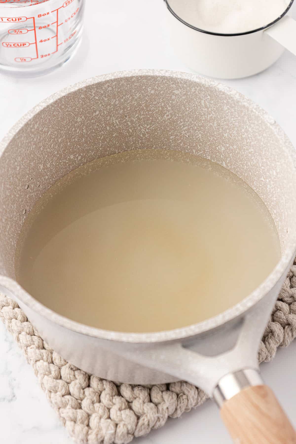 A beige saucepan with a light brown handle, filled with a cloudy liquid, sits on a woven trivet. In the background are a clear measuring cup with water and a white pot containing sugar.