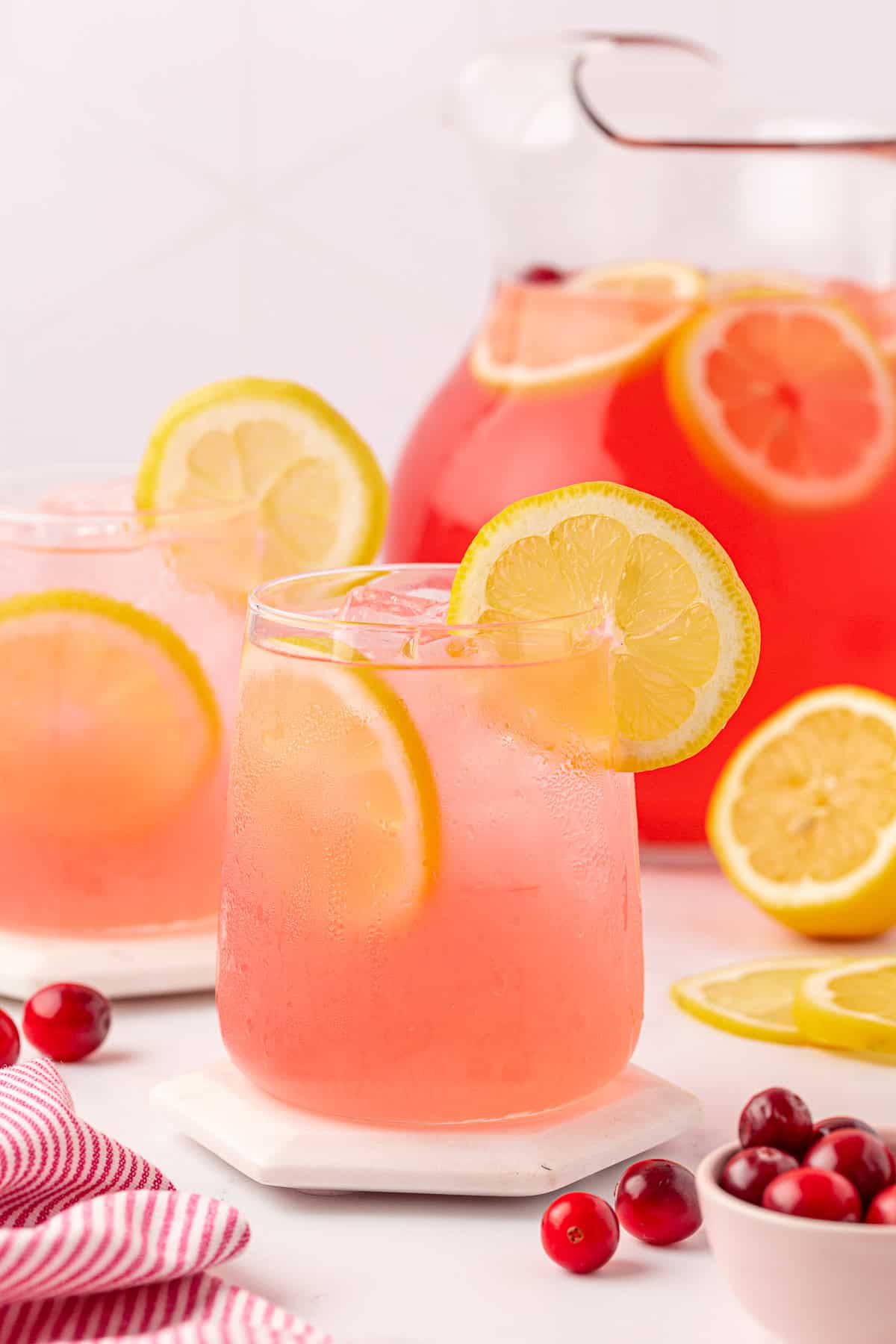 A glass of pink lemonade with ice and lemon slices, garnished with a lemon wheel, sits on a coaster. A pitcher of pink lemonade and extra lemon slices are in the background, with cranberries and a red-striped cloth nearby.