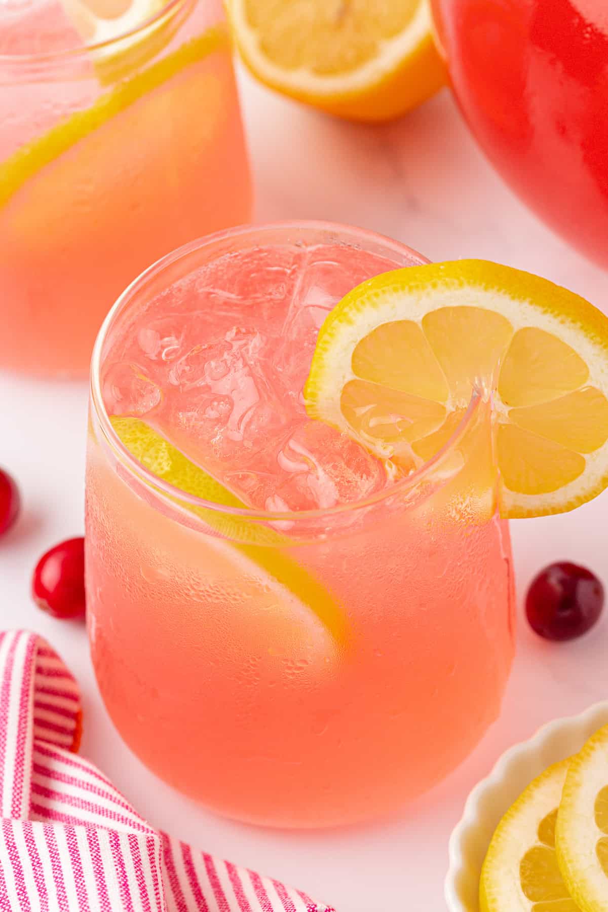 A glass of pink lemonade with ice, garnished with a lemon slice, sits on a white surface near fresh lemons, cherries, and a pink-striped cloth. Another glass and a pitcher are visible in the background.