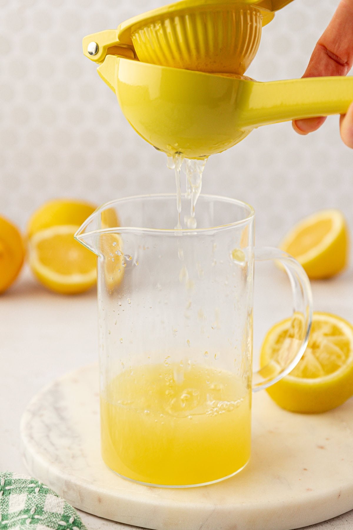 A hand squeezes a yellow citrus juicer over a glass pitcher, extracting fresh lemon juice. Halved lemons and a green towel are visible on a white surface in the background.