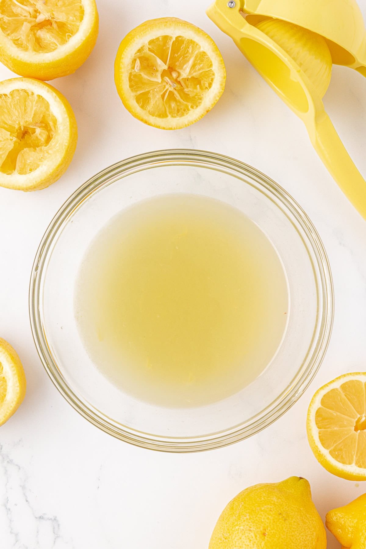 A glass bowl filled with lemon juice sits on a white surface, surrounded by halved and whole lemons, and a yellow lemon squeezer.
