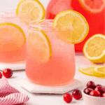 A glass of pink lemonade with ice and lemon slices, garnished with a lemon wheel. Cranberries and lemon slices are nearby, with another glass and a large pitcher in the background.