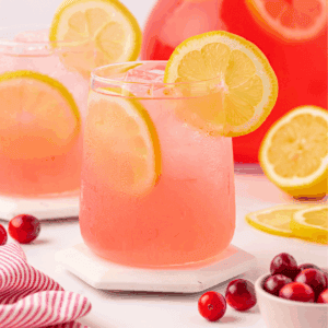 A glass of pink lemonade with ice and lemon slices, garnished with a lemon wheel. Cranberries and lemon slices are nearby, with another glass and a large pitcher in the background.