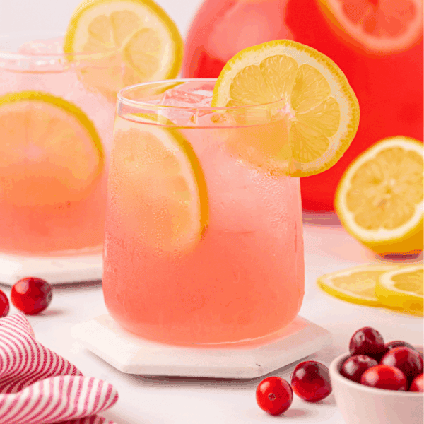 A glass of pink lemonade with ice and lemon slices, garnished with a lemon wheel. Cranberries and lemon slices are nearby, with another glass and a large pitcher in the background.