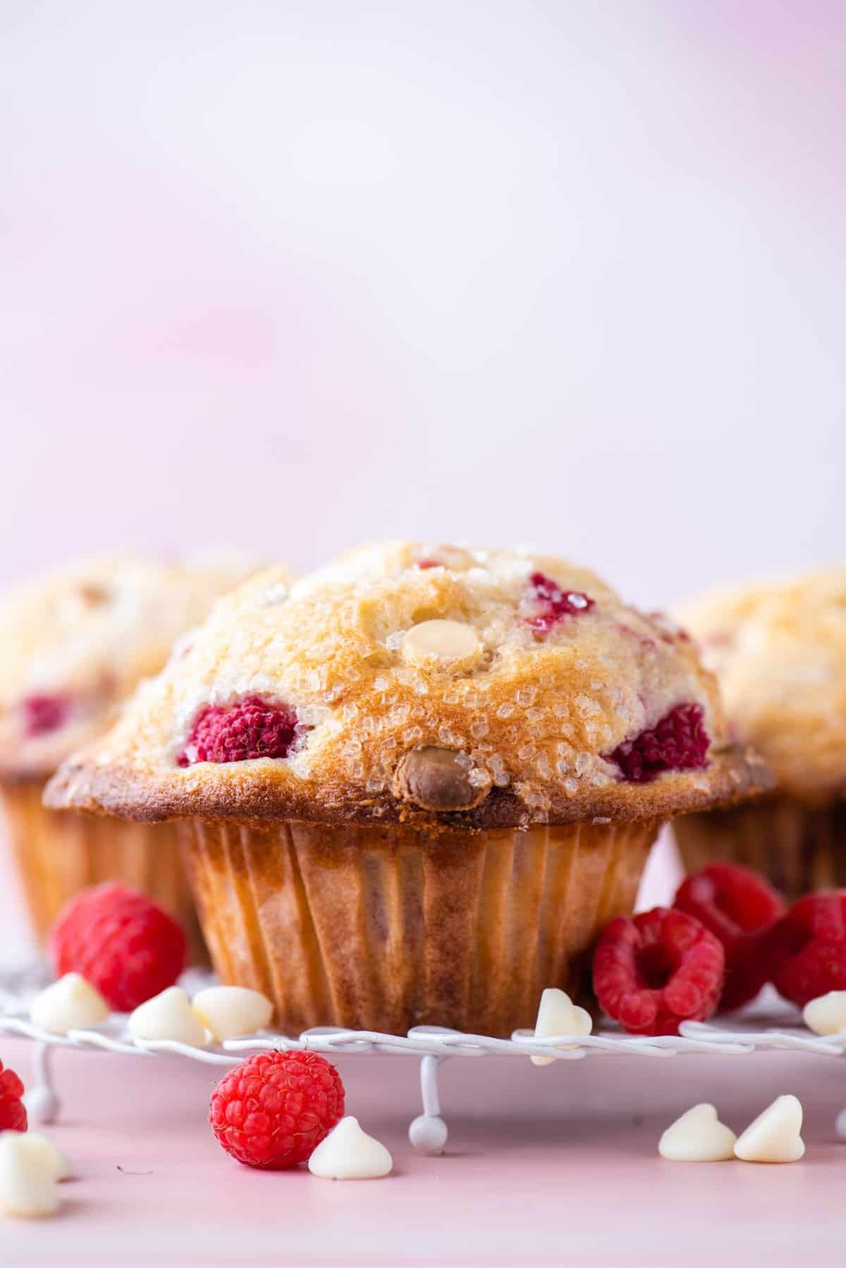 Close-up of a raspberry muffin topped with sugar crystals, surrounded by fresh raspberries and white chocolate chips on a white plate, with a soft pink background.