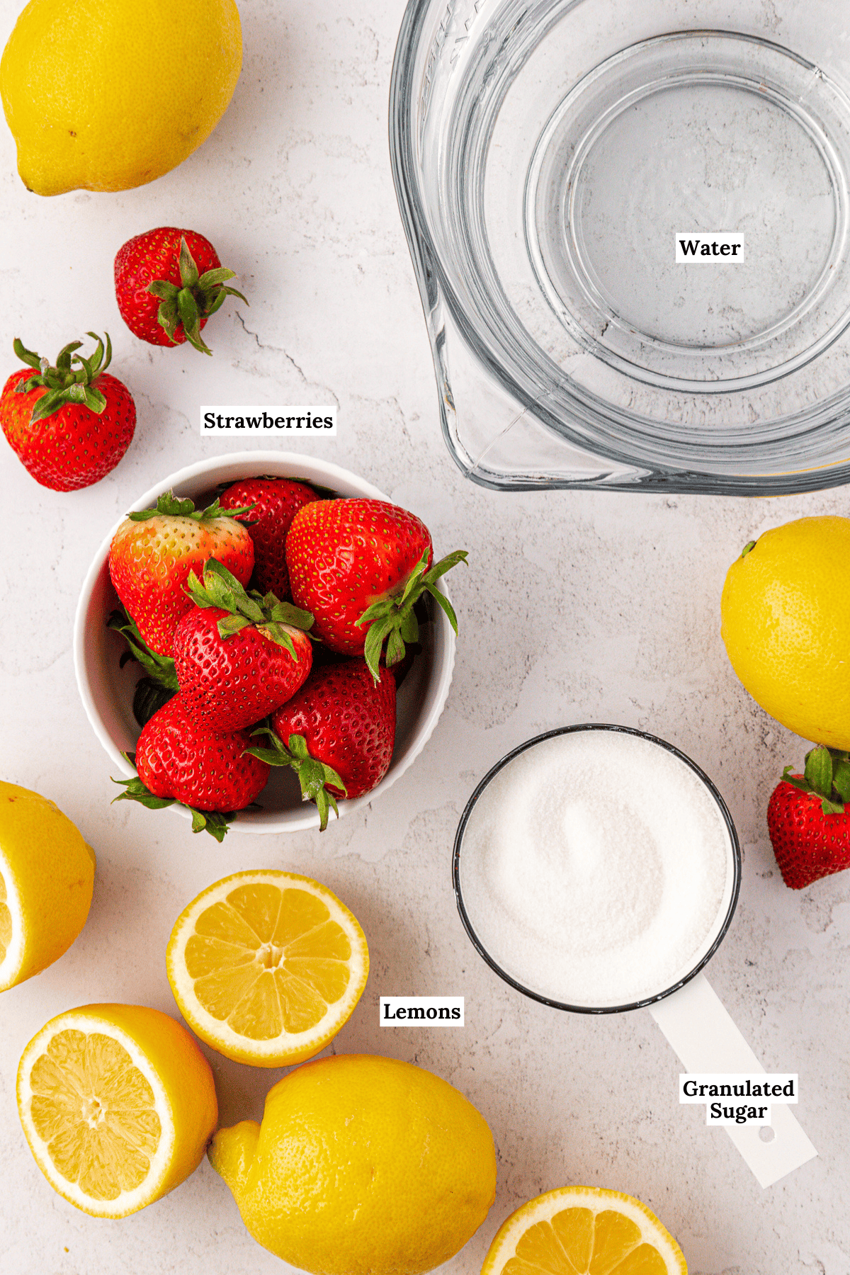 Top-down view of fresh strawberries in a bowl, whole and halved lemons, granulated sugar in a measuring cup, and a bowl of water on a white surface. Each ingredient is labeled: strawberries, lemons, sugar, and water.