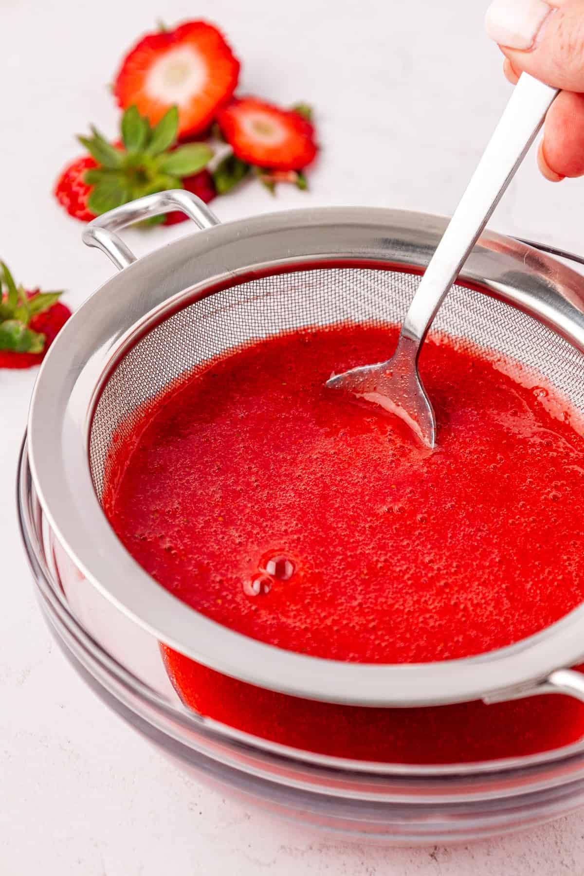 A hand uses a spoon to strain fresh strawberry puree through a fine mesh sieve into a clear glass bowl, with sliced strawberries and green leaves in the background.