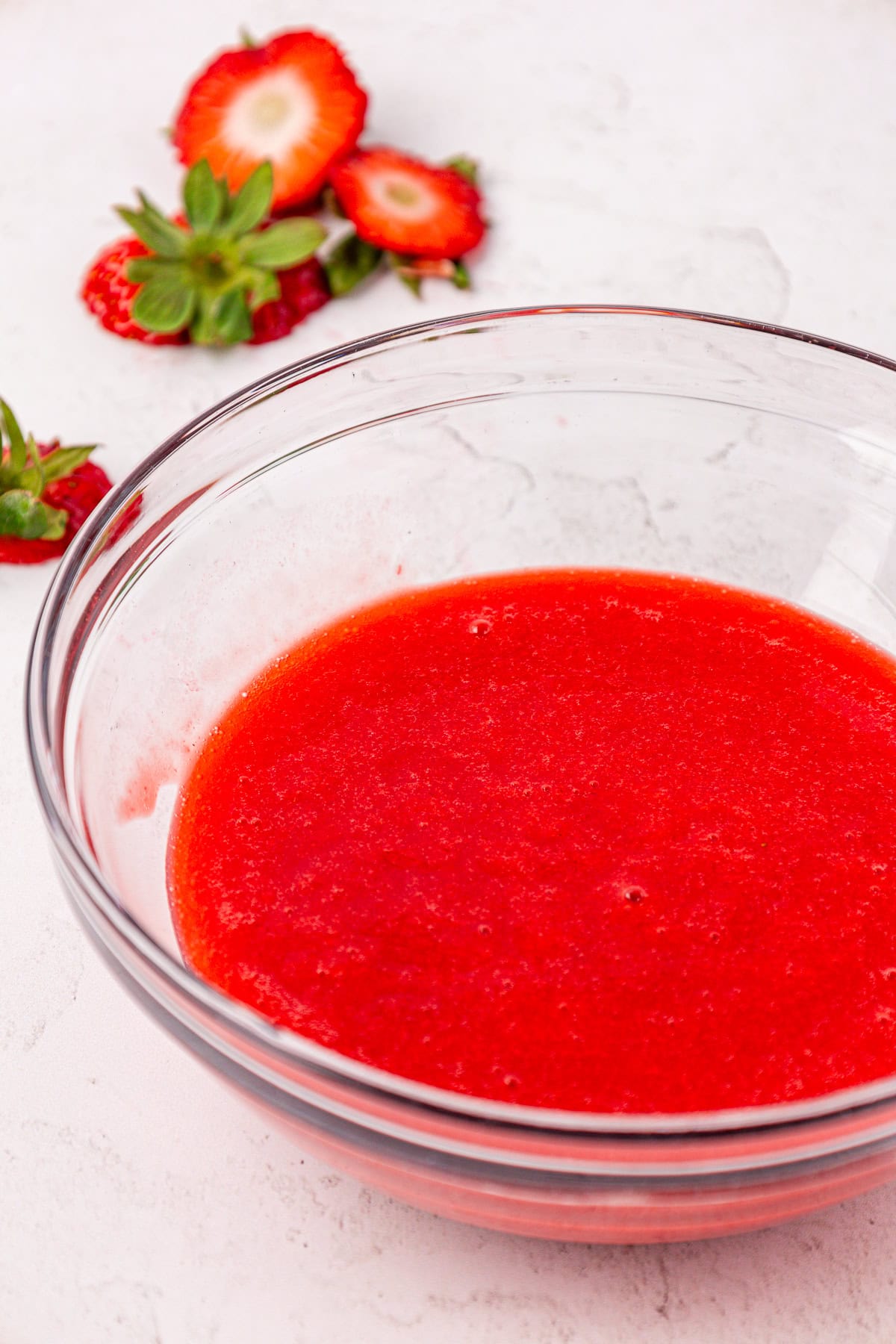 A clear glass bowl filled with bright red strawberry puree sits on a light surface, with fresh strawberry slices and whole strawberries in the background.