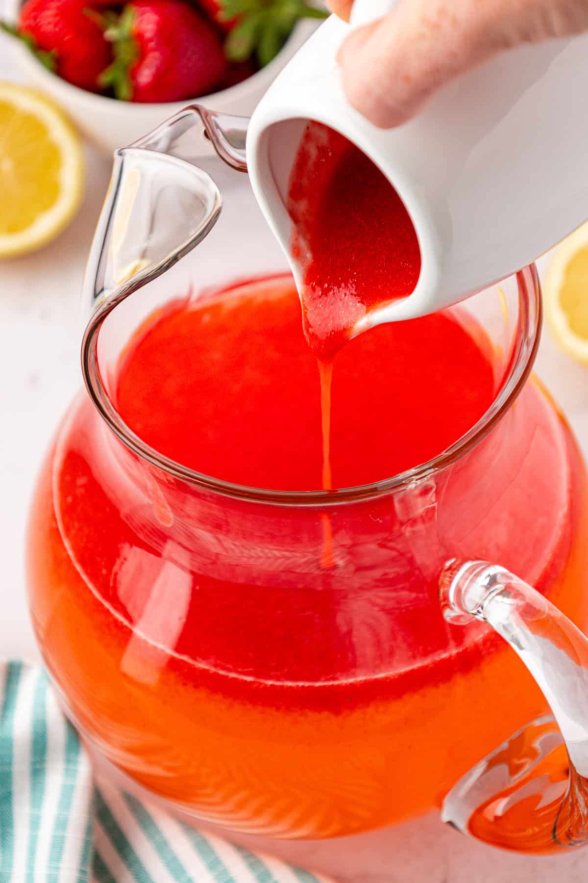 A hand pours bright red strawberry puree from a white pitcher into a clear glass jug filled with orange-red liquid, with strawberries, lemon slices, and a striped towel nearby.