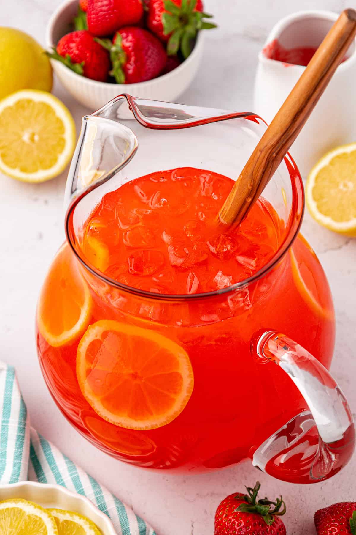A glass pitcher filled with iced strawberry lemonade, garnished with lemon slices, sits on a table surrounded by fresh strawberries and lemon halves. A wooden spoon rests inside the pitcher.