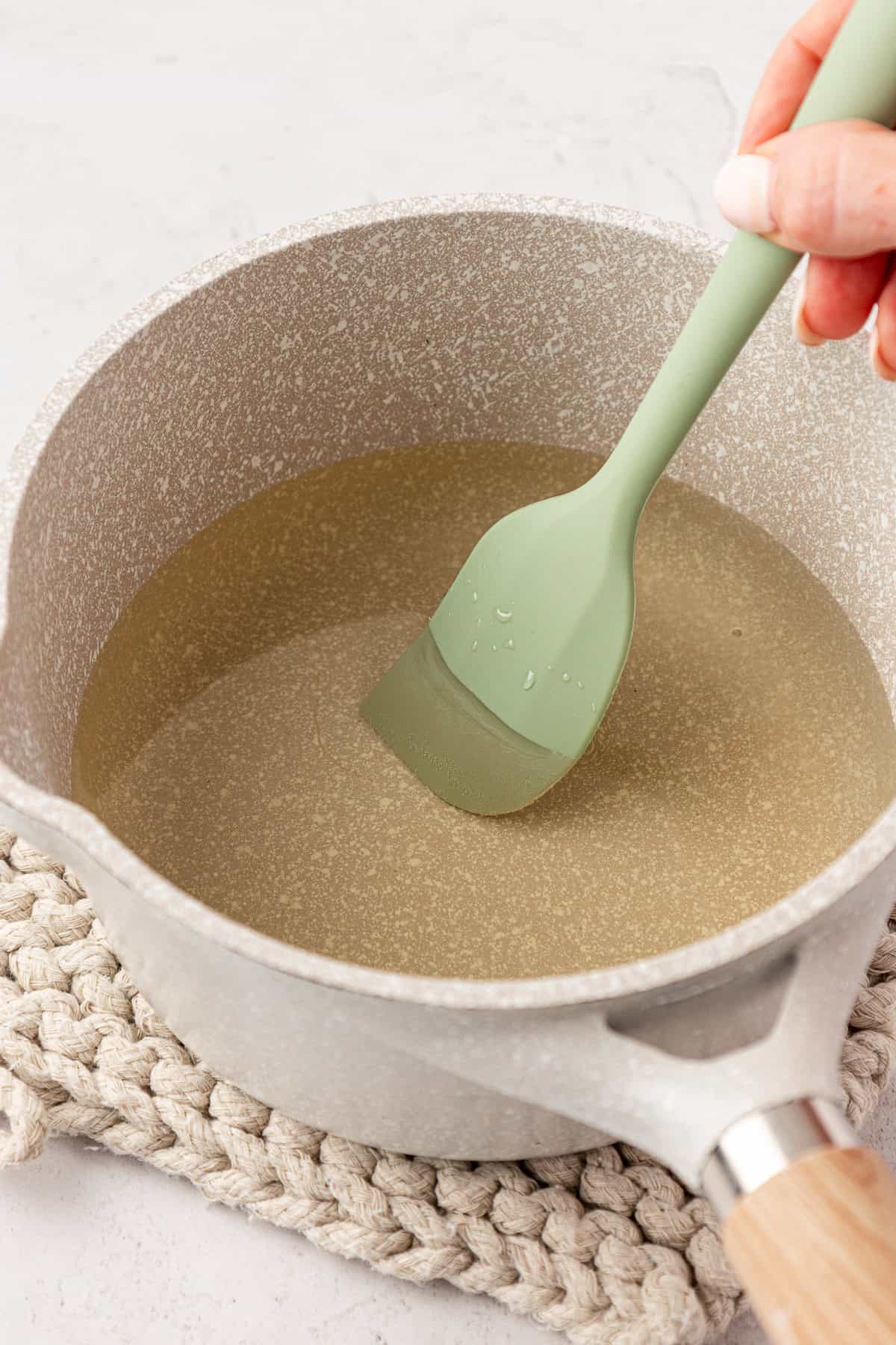 A hand stirs a clear liquid in a gray saucepan with a green silicone spatula, placed on a beige woven mat.