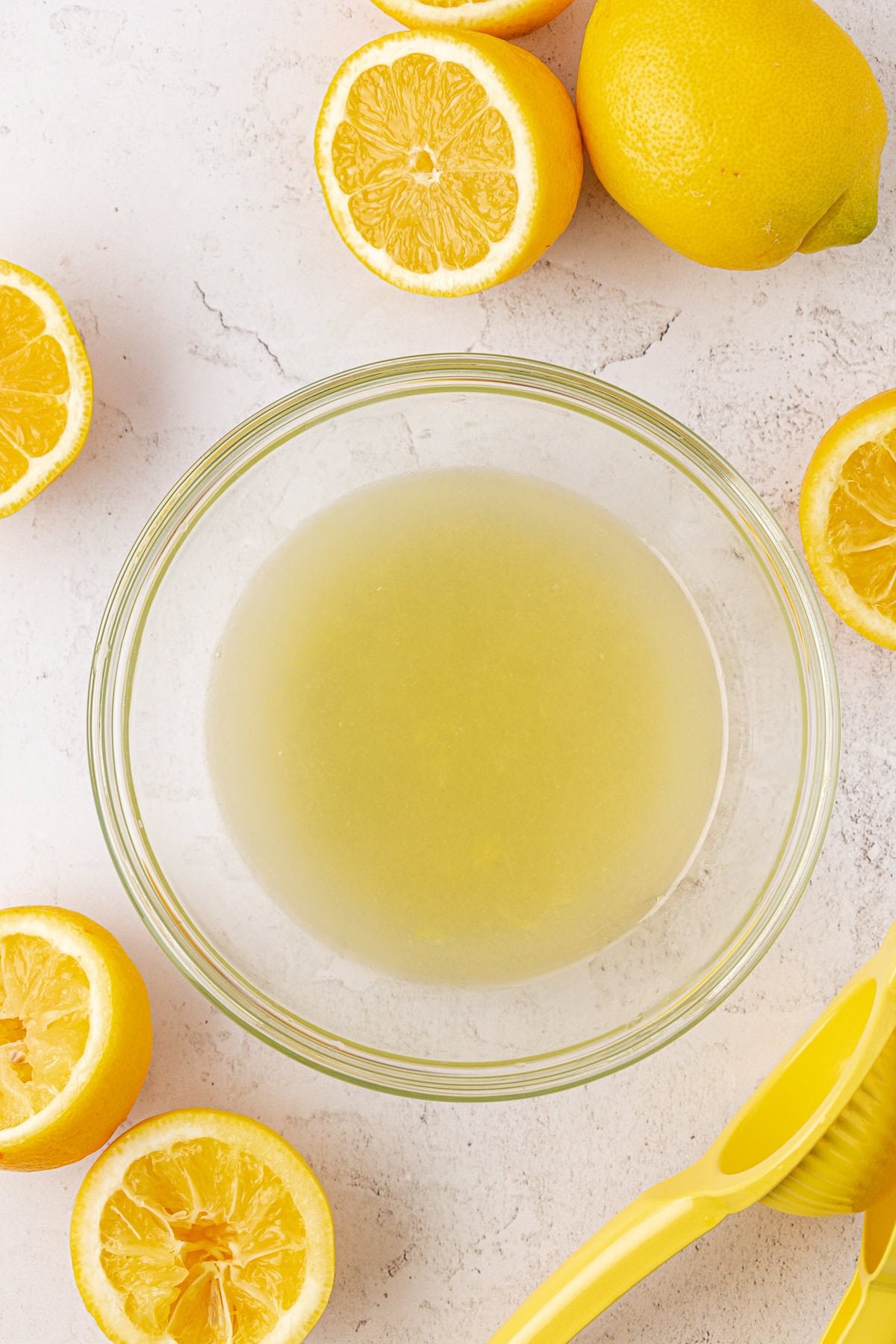 A glass bowl filled with lemon juice sits on a light surface, surrounded by several halved and whole lemons, with a yellow citrus juicer nearby.
