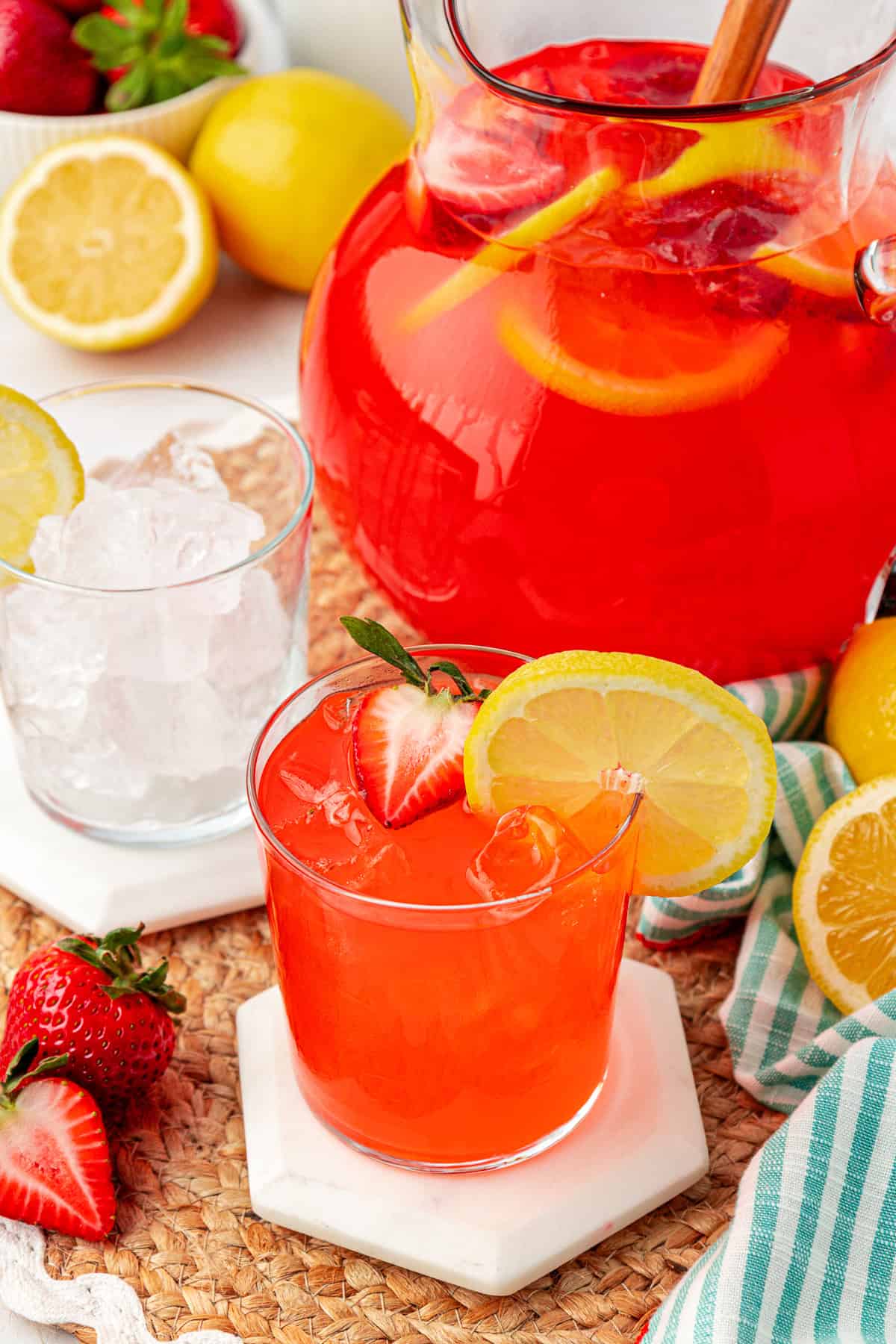 A glass of bright red strawberry lemonade with ice, garnished with a lemon slice and strawberry, sits on a coaster next to a pitcher of the drink. Sliced lemons, strawberries, and a glass of ice are nearby.