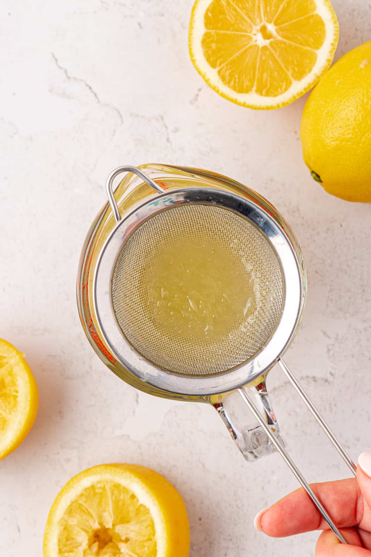 A hand holds a metal strainer over a glass jar, filtering lemon juice. Halved and whole lemons are scattered on a white surface around the jar.