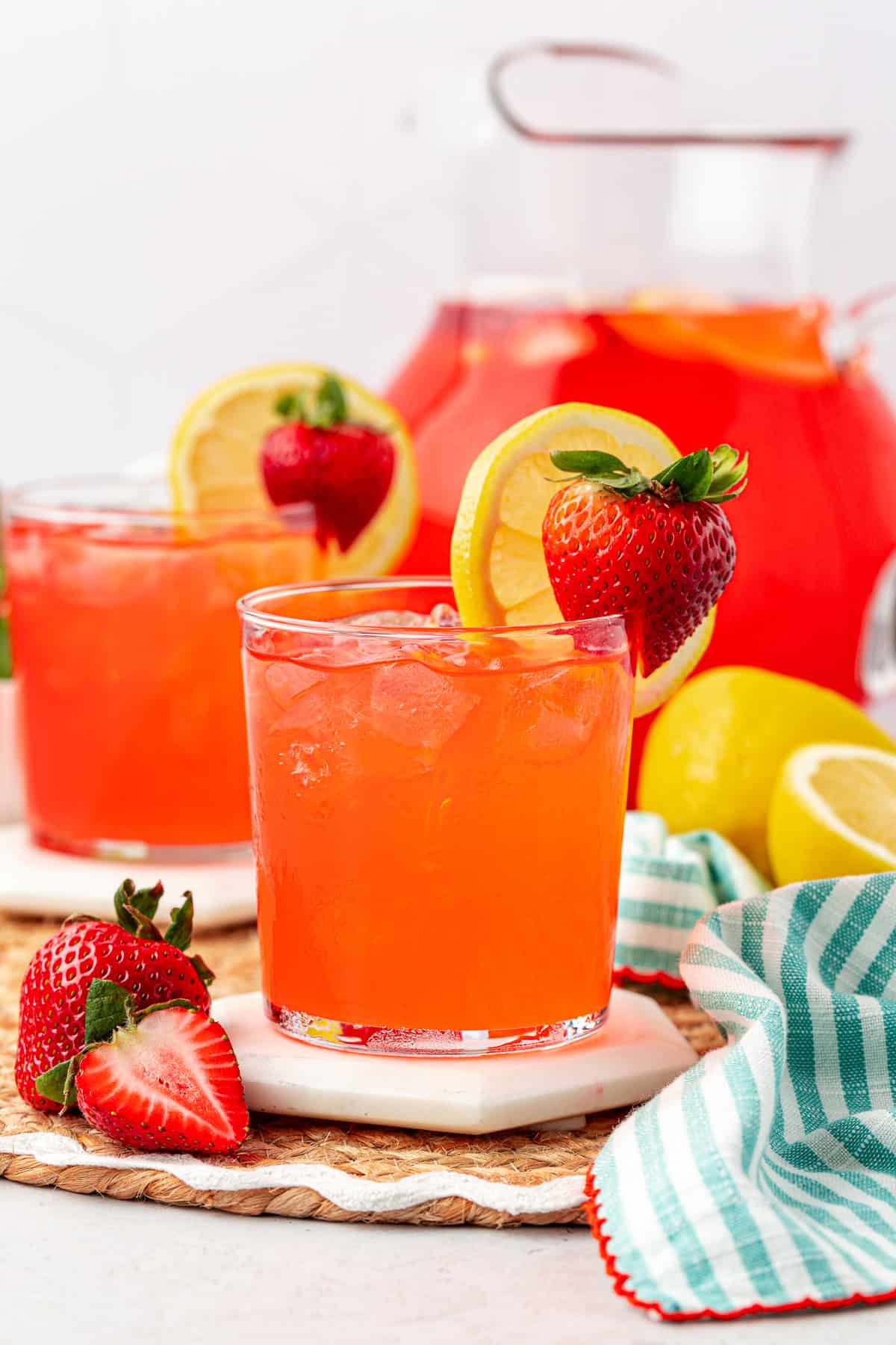 Two glasses of strawberry lemonade with ice, garnished with lemon slices and whole strawberries, sit on coasters. A pitcher of the drink, fresh strawberries, lemons, and a striped cloth are nearby.