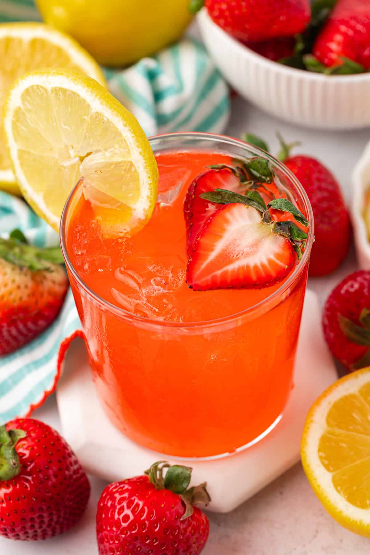 A glass of bright red strawberry lemonade with ice, garnished with a lemon slice and a halved strawberry, surrounded by fresh strawberries and a striped cloth in the background.