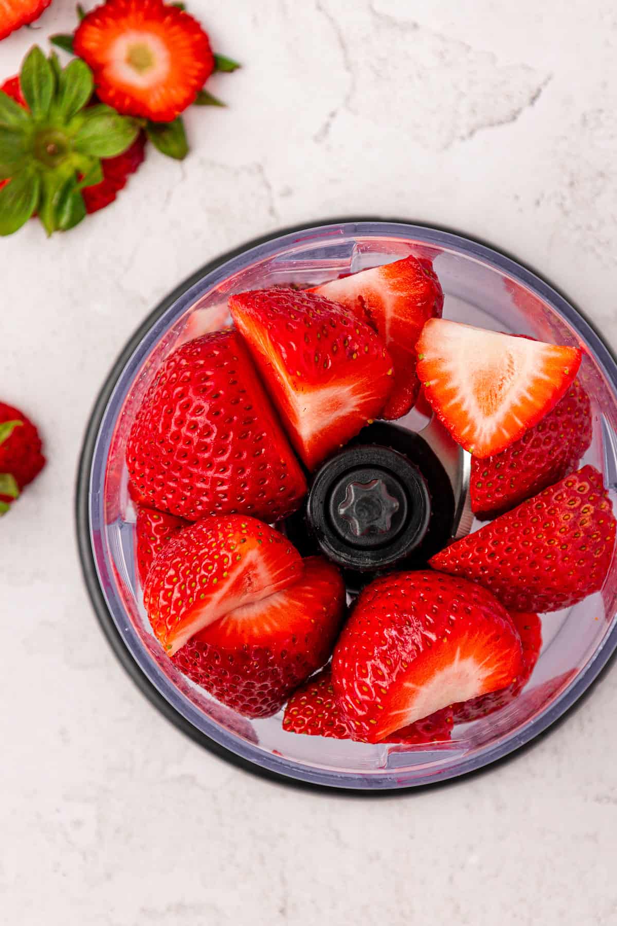Fresh strawberries, some halved, are arranged inside a small food processor on a light-colored surface, with whole strawberries and green leaves nearby.