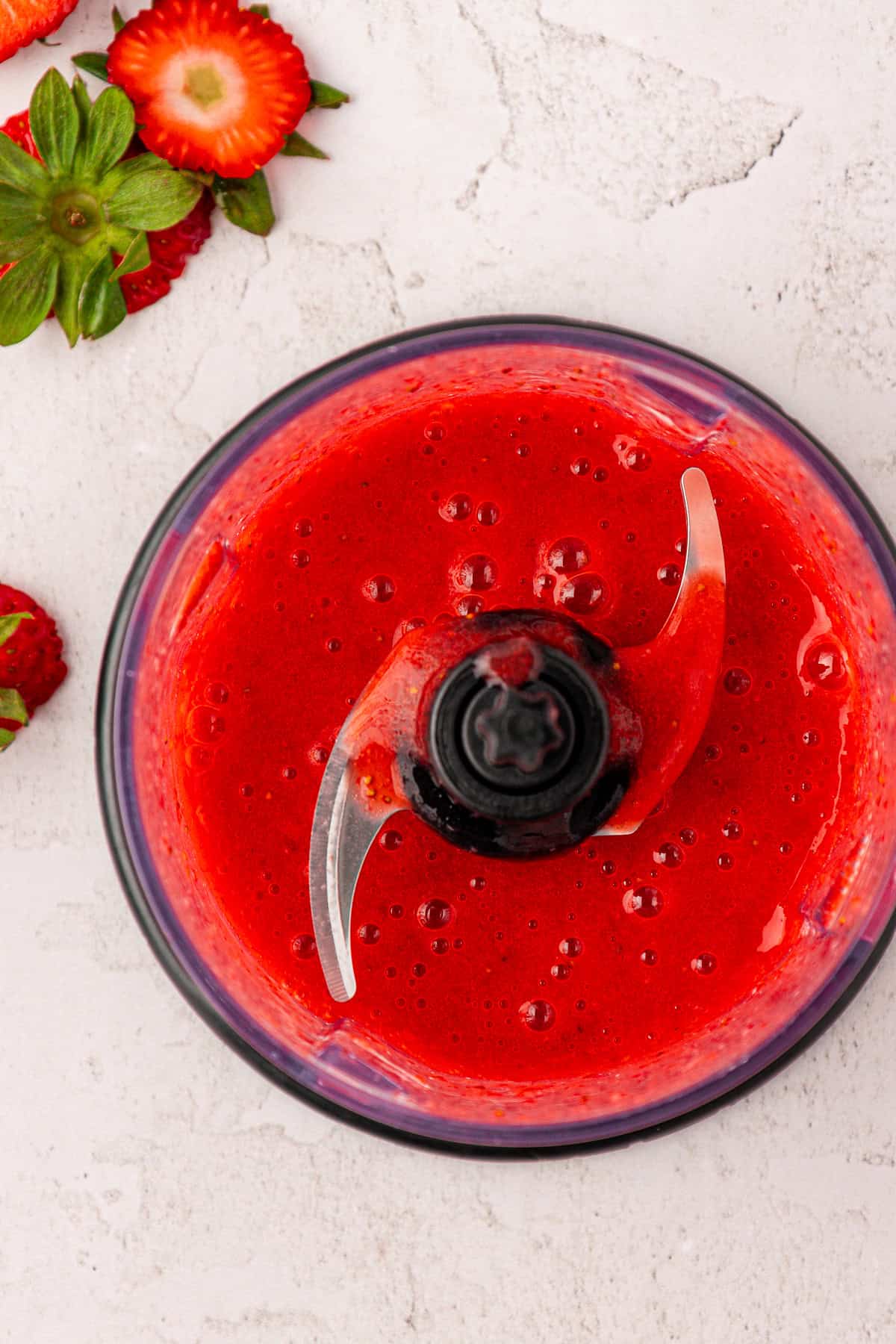 A food processor bowl filled with smooth, bright red strawberry puree. Whole and sliced strawberries are scattered on a light-colored surface next to the bowl.
