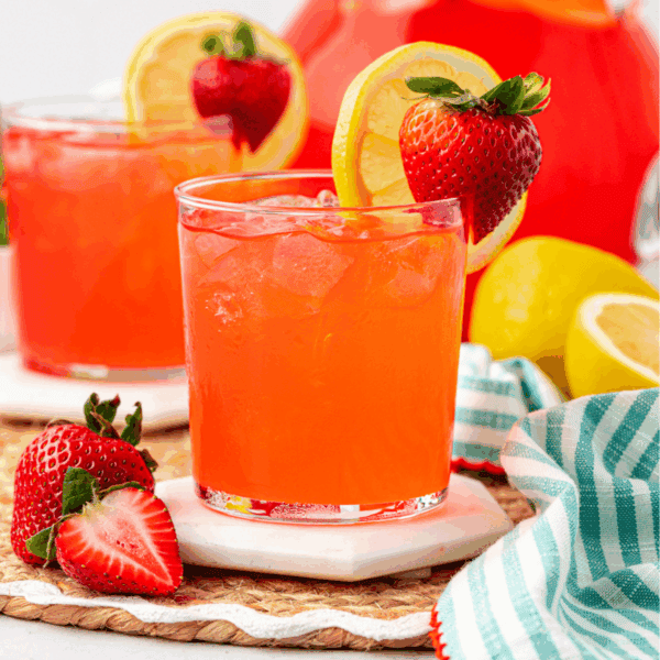 A glass of bright red strawberry lemonade with ice, garnished with a lemon slice and strawberry. Another drink, strawberries, lemons, and a striped cloth are in the background on a woven mat.