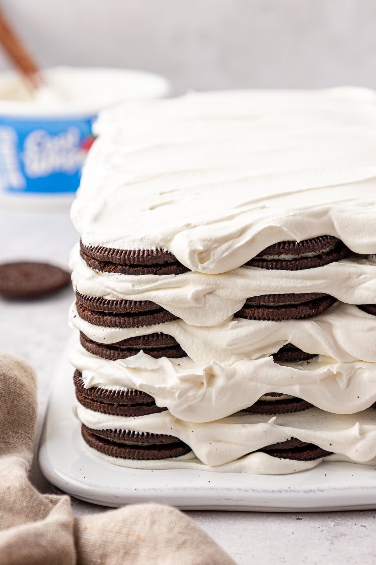 A close-up of a layered dessert made with chocolate sandwich cookies and whipped cream, forming a rectangular cake shape on a white surface. A container of whipped topping and a cookie are in the background.