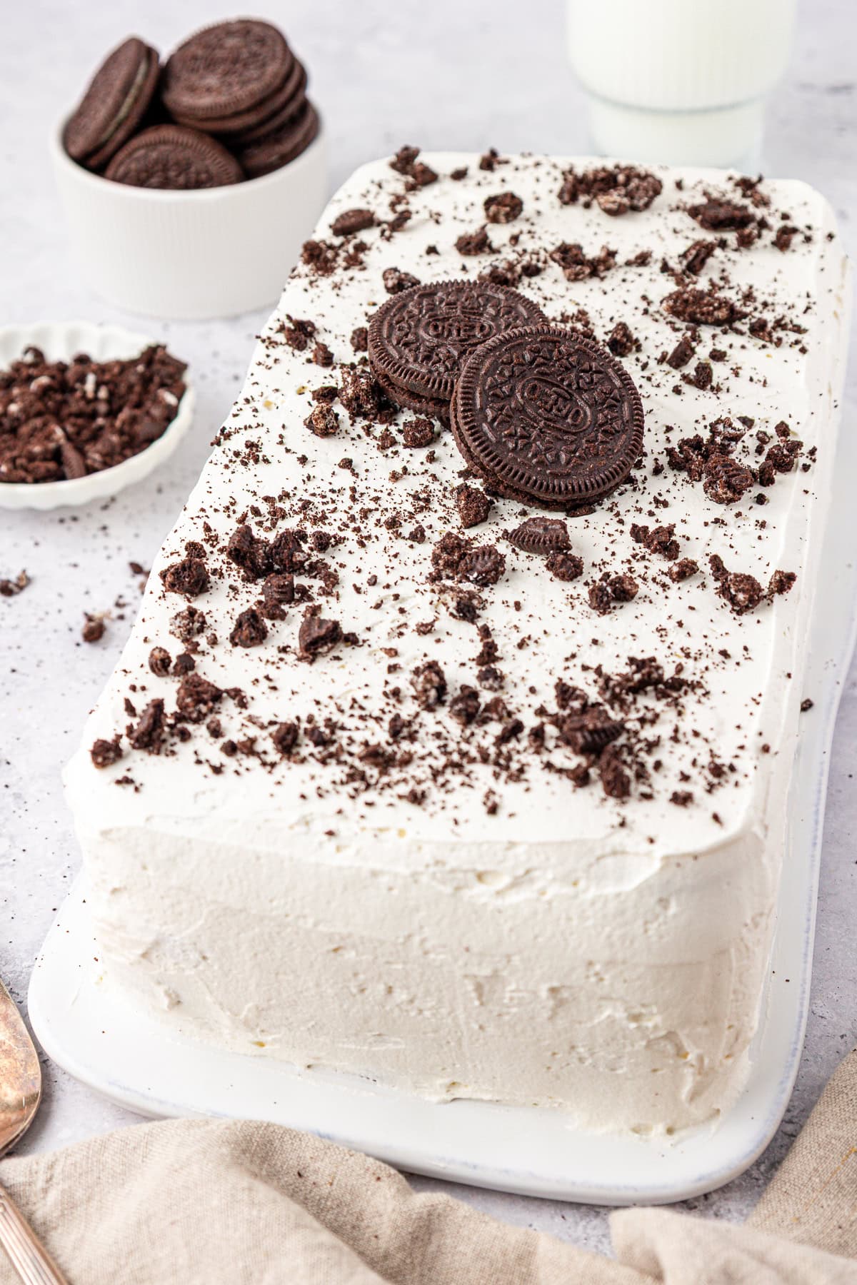 Rectangular cookies and cream ice cream cake topped with whole and crushed chocolate sandwich cookies, placed on a white platter. Bowls of cookies and cookie crumbs are in the background.