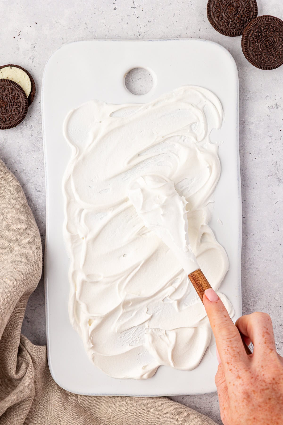 A hand spreads creamy white frosting on a rectangular white cutting board with a wooden-handled spatula. Chocolate sandwich cookies and a beige cloth are nearby on a light surface.