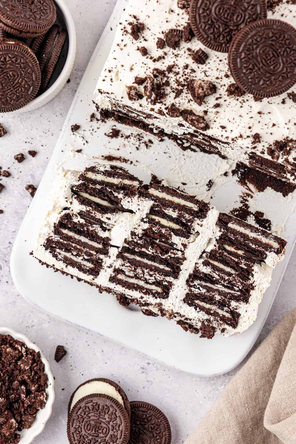 A sliced Oreo icebox cake on a white platter shows layers of chocolate cookies and whipped cream, with whole and crushed Oreo cookies scattered nearby on a light countertop.