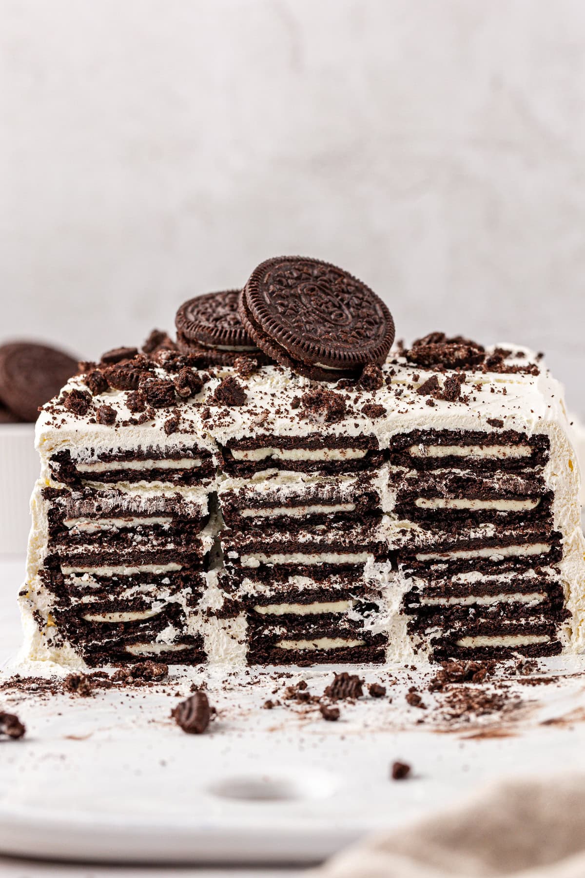A layered cookies and cream icebox cake with visible rows of chocolate sandwich cookies and cream, topped with more cookies and crumbled pieces, on a white surface.