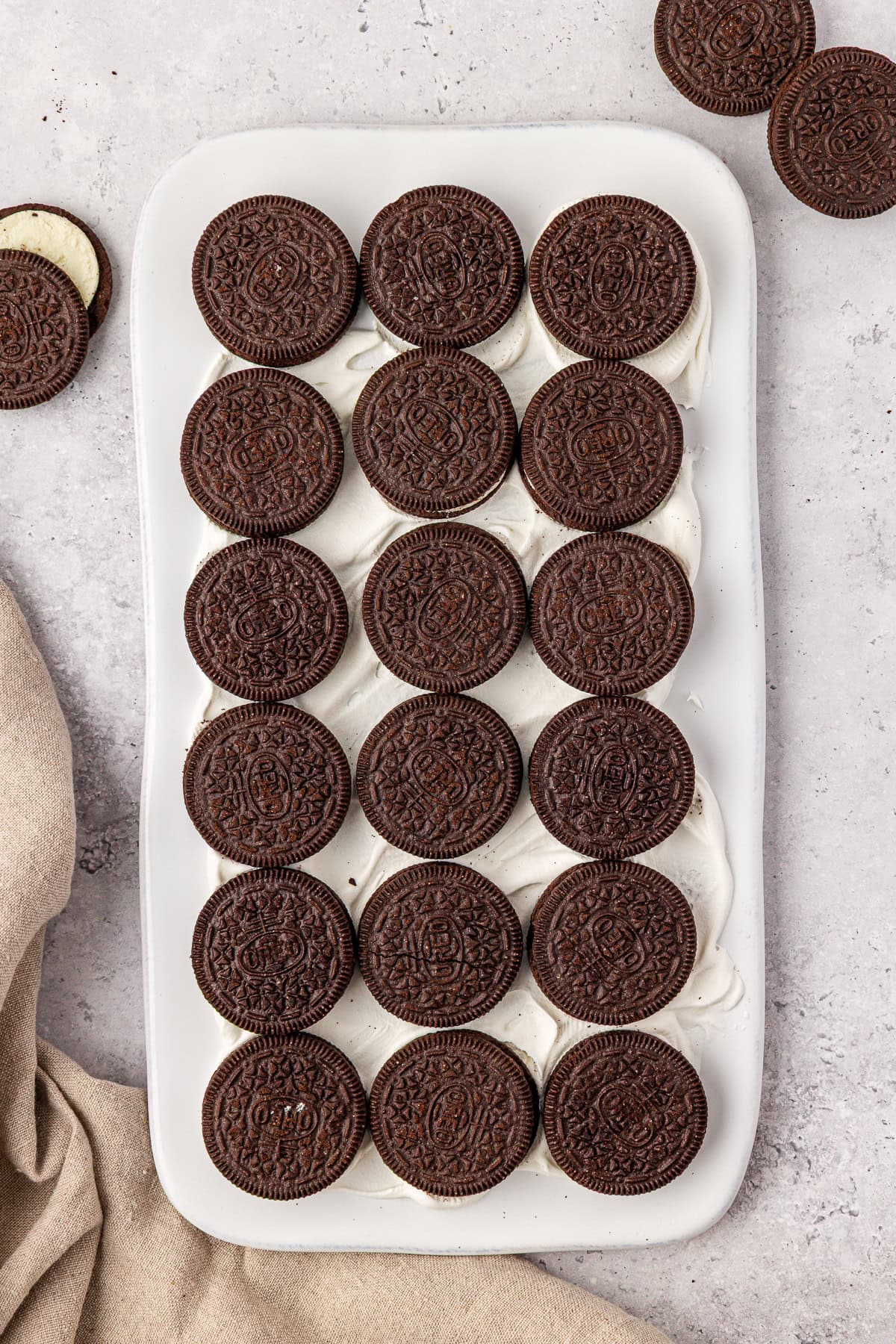 A white rectangular dish filled with rows of chocolate sandwich cookies arranged neatly on top of a layer of creamy white filling, set on a light gray countertop with a beige cloth nearby.