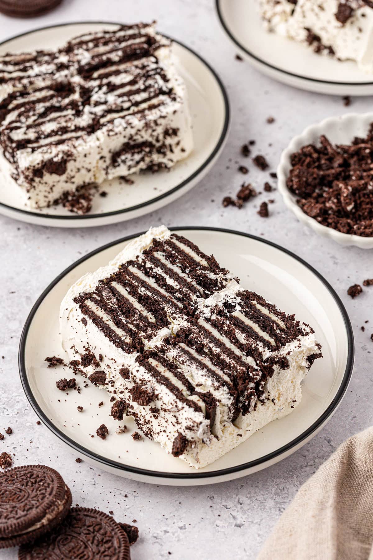 Two slices of cookies and cream icebox cake are served on a plate, featuring layers of chocolate cookies and whipped cream. Crumbled cookies are scattered around, with extra plates and whole cookies in the background.