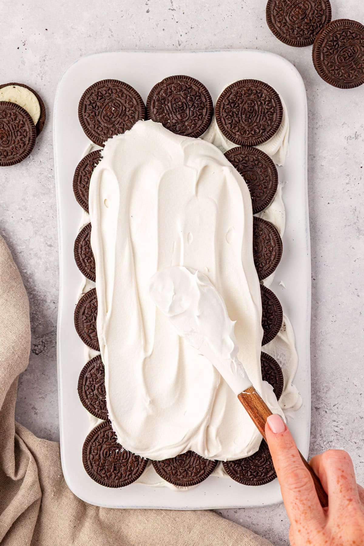 A hand spreads whipped cream over a layer of chocolate sandwich cookies arranged in a white rectangular dish, with extra cookies and a beige cloth nearby on a light surface.