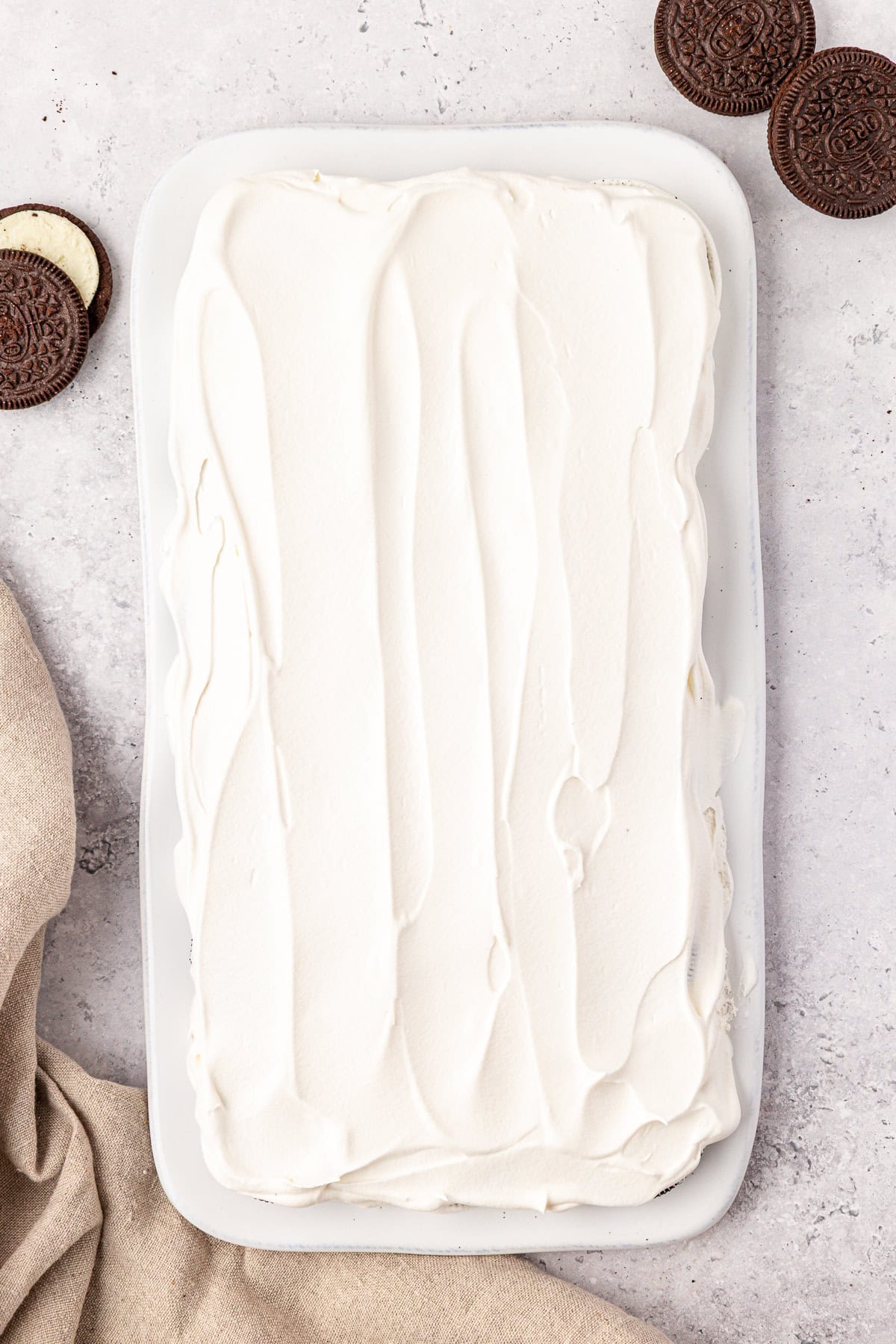 A rectangular dish filled with a dessert covered in smooth, white whipped topping. Chocolate sandwich cookies are placed nearby on a light textured surface, with a beige cloth partially visible in the corner.
