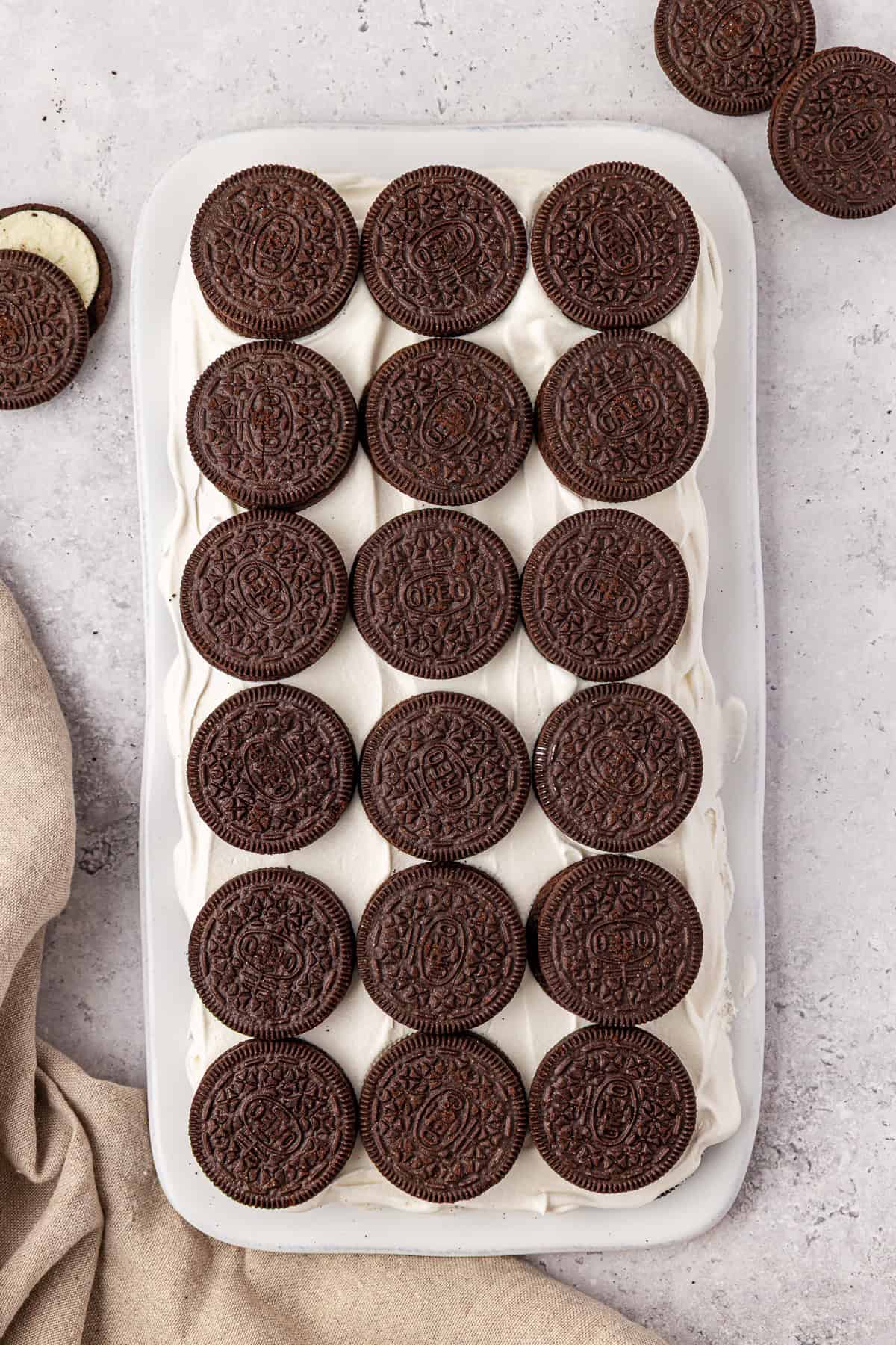 A rectangular tray of dessert topped with neatly arranged whole chocolate sandwich cookies, placed on a light surface with a beige cloth and a few extra cookies nearby.