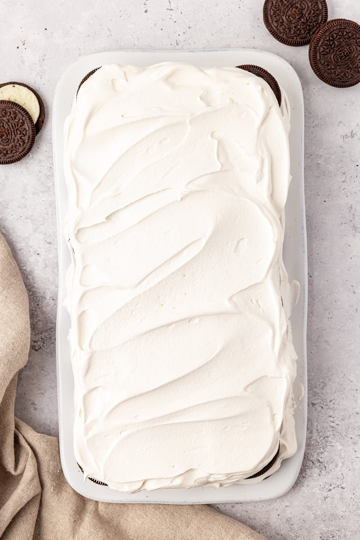 A rectangular dessert topped with smooth white whipped cream sits on a white tray, with chocolate sandwich cookies and a beige cloth beside it on a light marble surface.