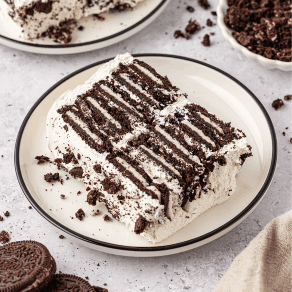 A slice of icebox cake with layers of chocolate cookies and whipped cream sits on a plate, with cookie crumbs scattered around and whole cookies visible nearby.