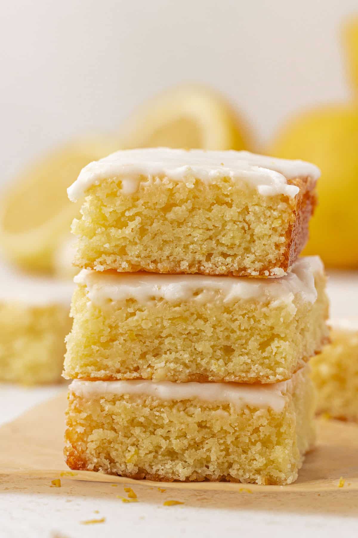 A stack of three lemon brownies with a light glaze on top, placed on parchment paper. The bars are moist, dense, and golden with a slightly crumbly texture. Lemons are blurred in the background.