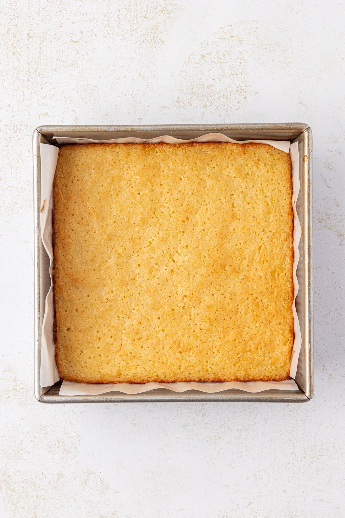 A golden-brown, freshly baked square cake sits in a parchment-lined metal baking pan on a light, textured surface.