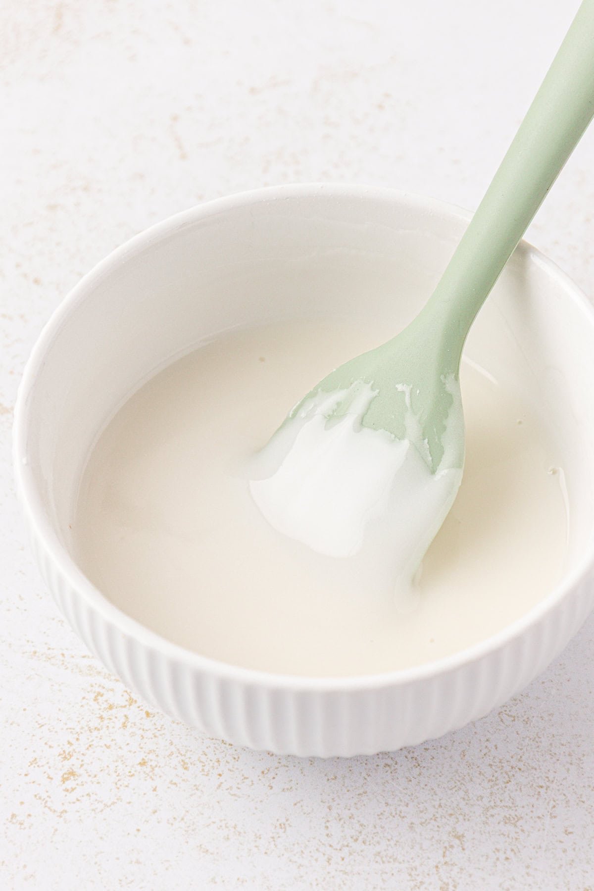 A white bowl filled with smooth white icing, being stirred with a light green silicone spatula, on a light background.