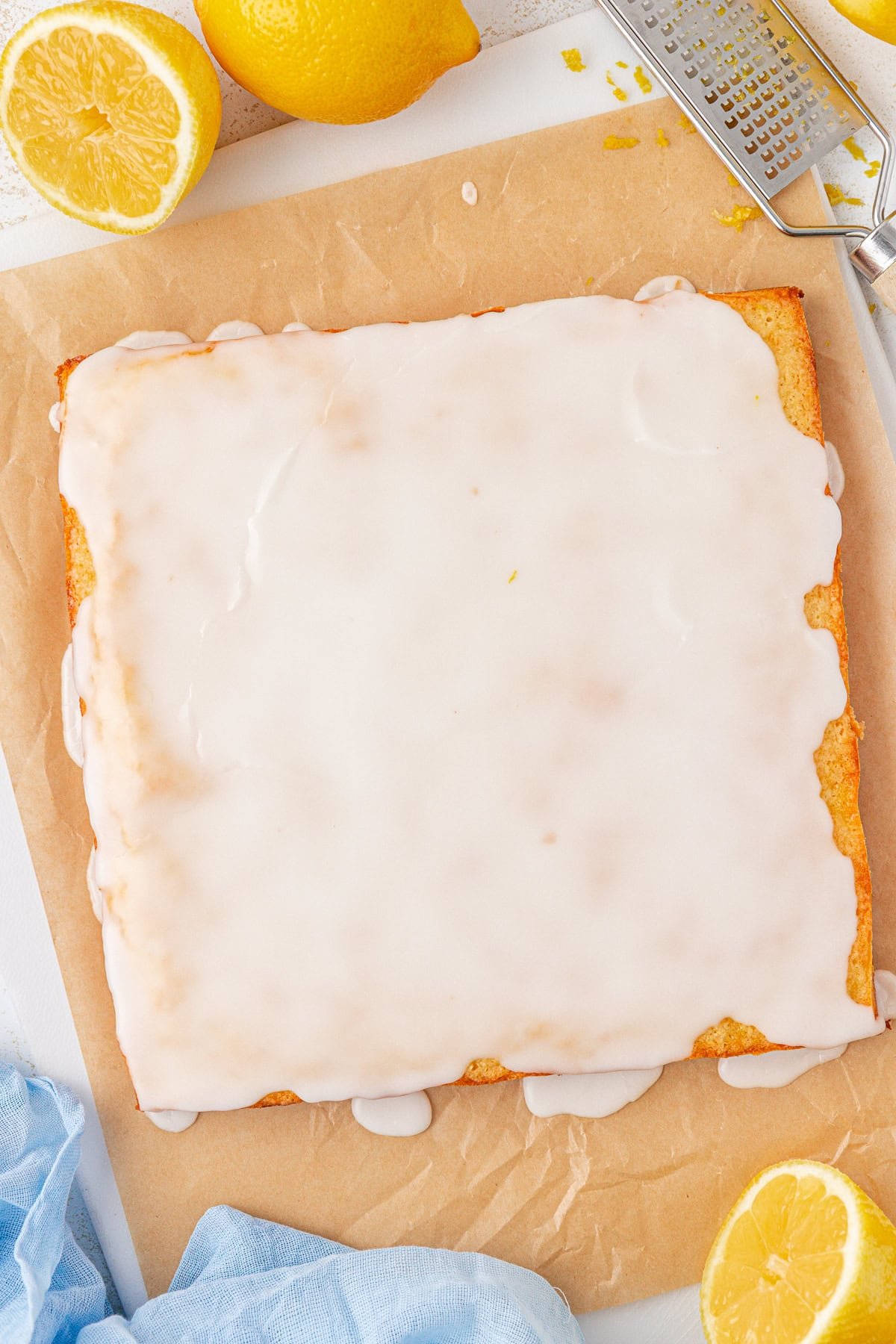 A rectangular lemon cake with white glaze sits on parchment paper, surrounded by fresh lemons and a grater. Some glaze drips down the sides of the cake. A blue cloth is partially visible in the corner.