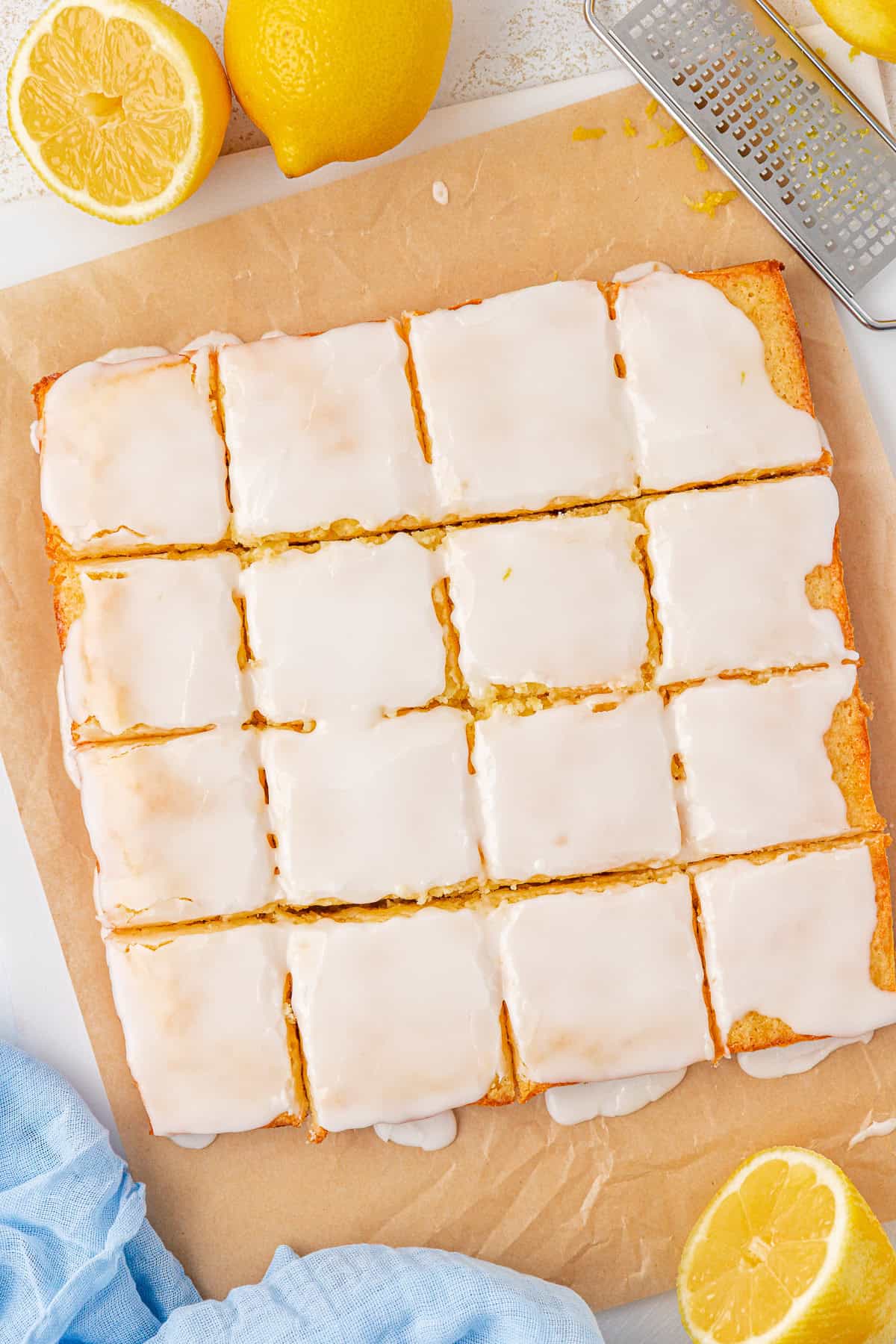 A batch of lemon brownies with white icing is cut into squares on parchment paper. Whole and halved lemons, a zester, and a light blue cloth are nearby on the white surface.