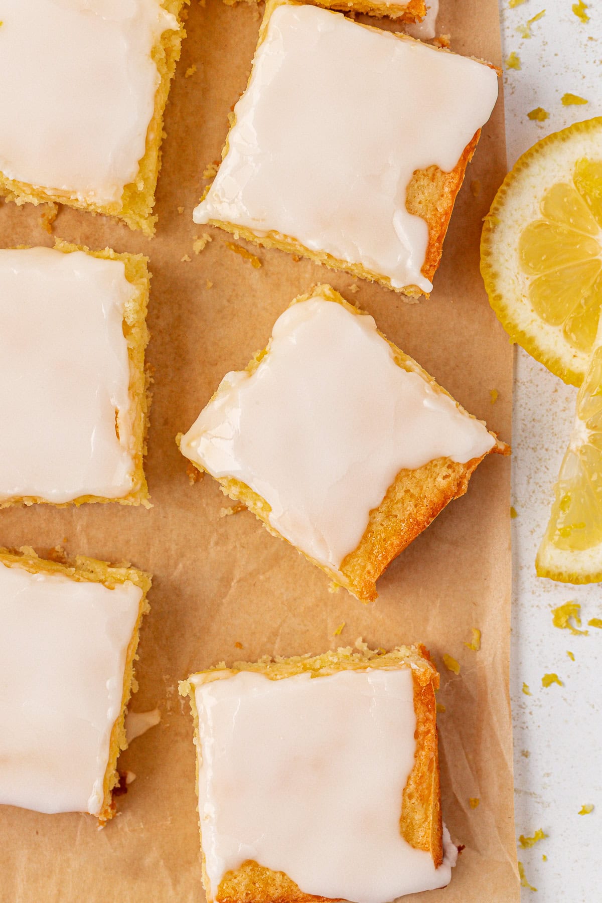 Several lemon brownies with glossy white icing are arranged on brown parchment paper, with a couple of fresh lemon slices nearby. The bars have a light, golden color and look soft and moist.