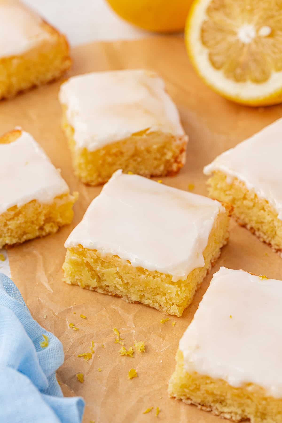 Six square lemon brownies with glossy white icing are arranged on brown parchment paper. A blue napkin and a halved lemon are nearby, adding a fresh, bright touch to the scene.