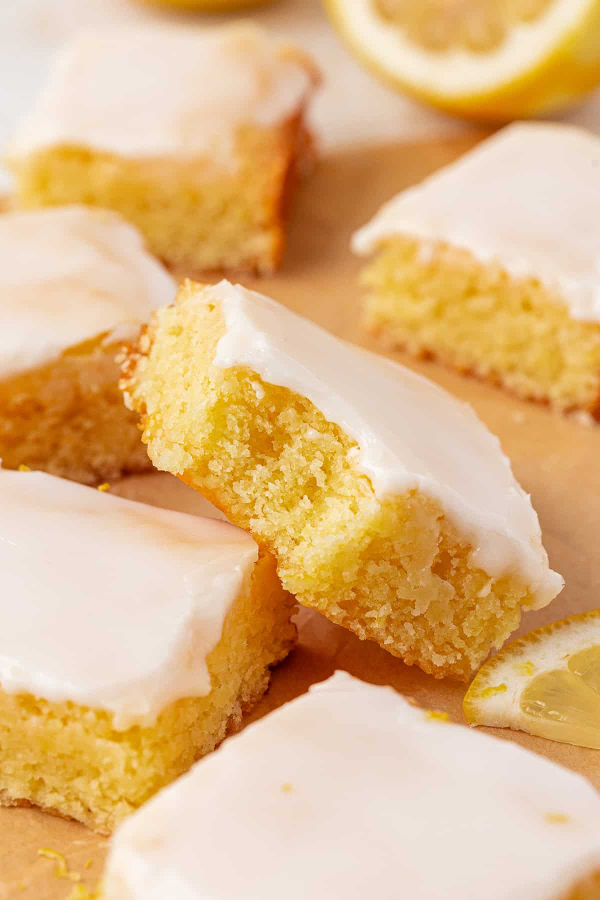 Close-up of glazed lemon bars with a moist, yellow cake texture and a glossy white icing on top, arranged on parchment paper with a lemon slice nearby.