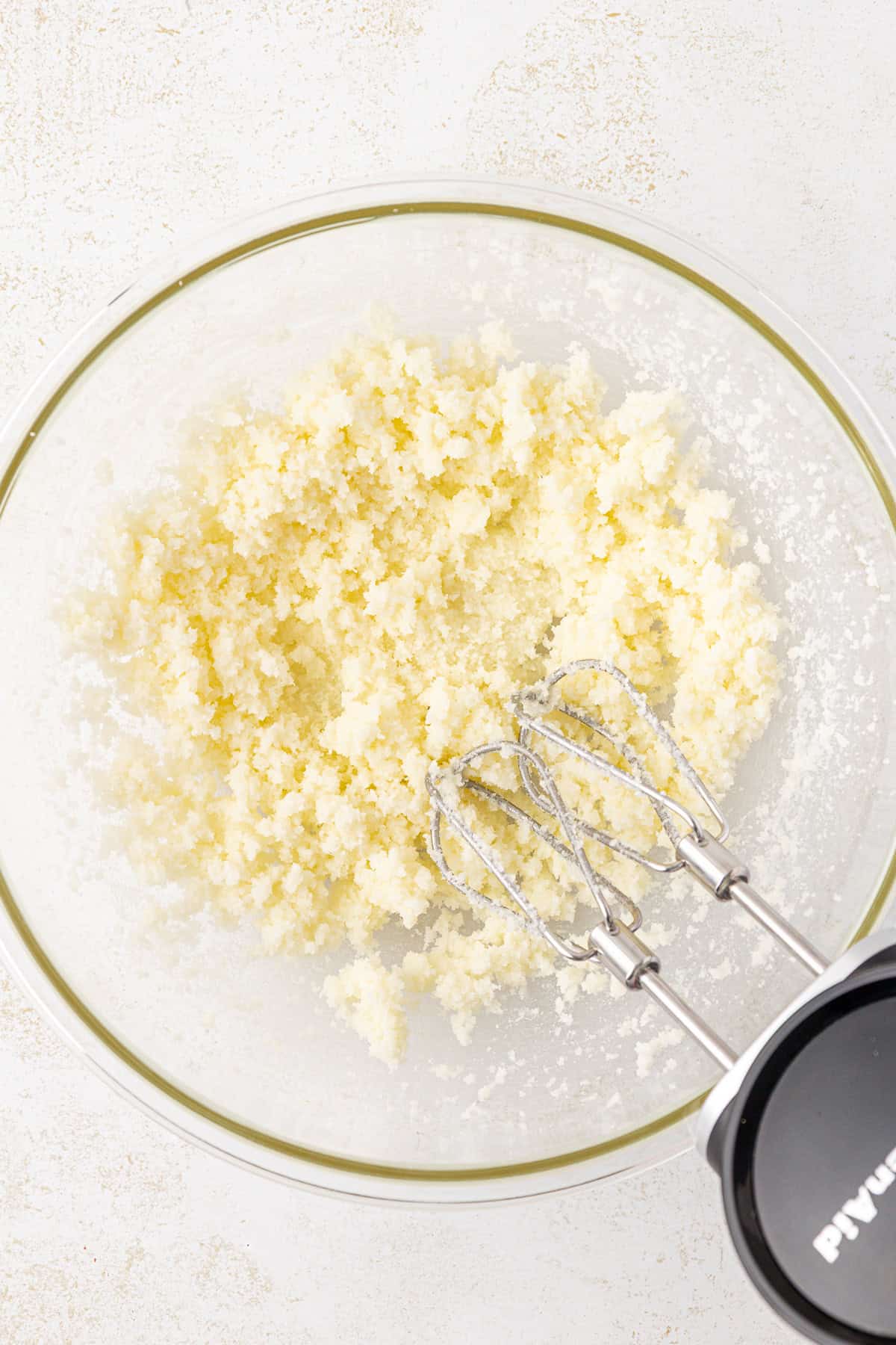 A glass bowl with creamed butter and sugar being mixed by a handheld electric mixer on a light, textured surface.