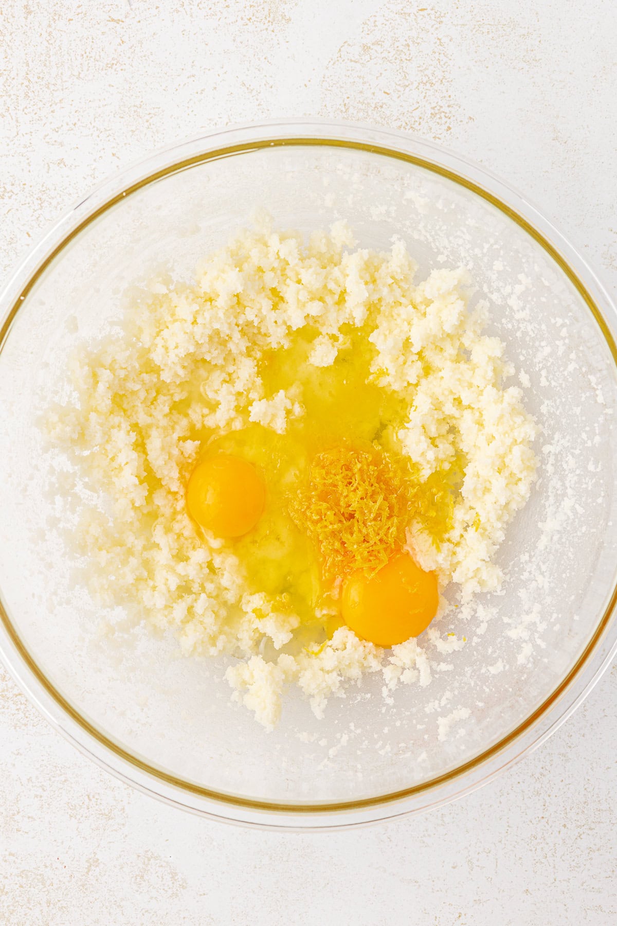 A glass bowl containing creamed butter and sugar, two raw eggs, and grated lemon zest, set on a white surface.