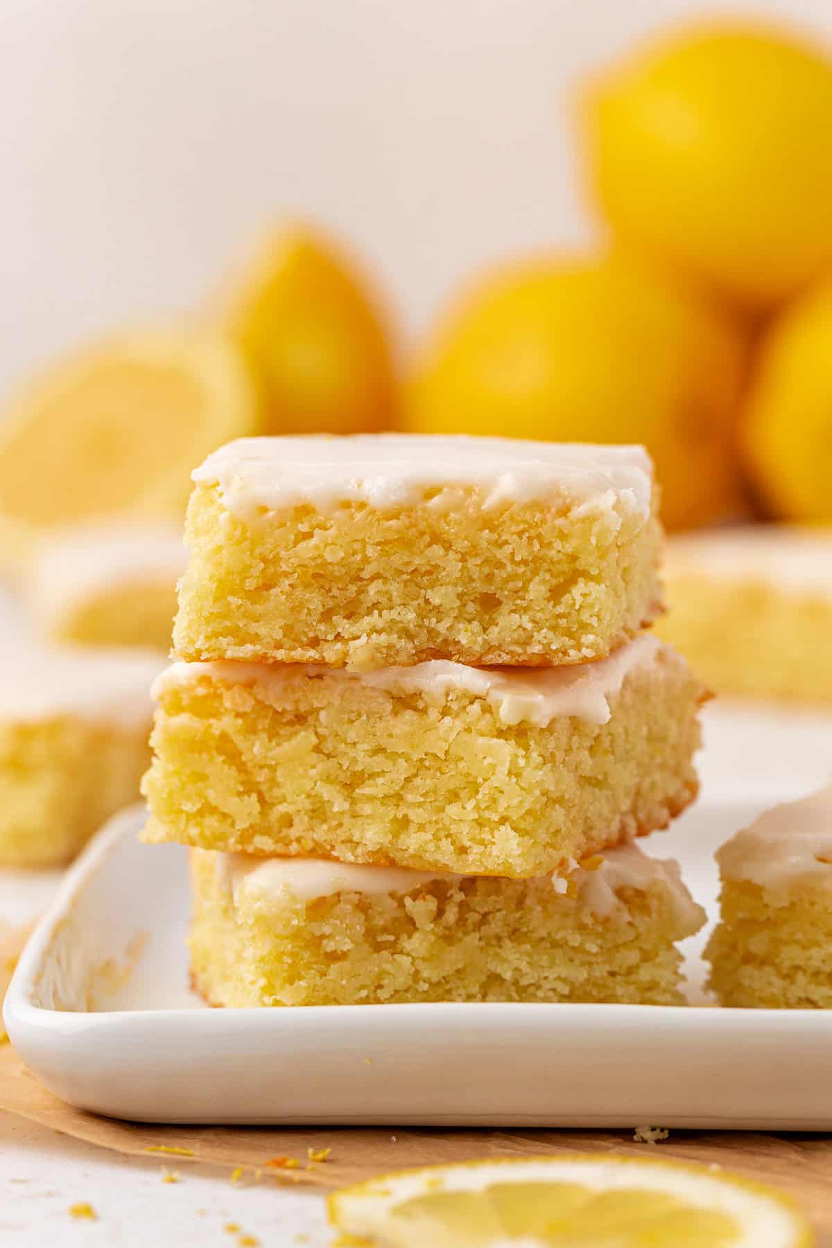 A stack of three lemon blondies with glossy icing on top, sitting on a white plate. Whole and sliced lemons are blurred in the background, suggesting a fresh lemon flavor.