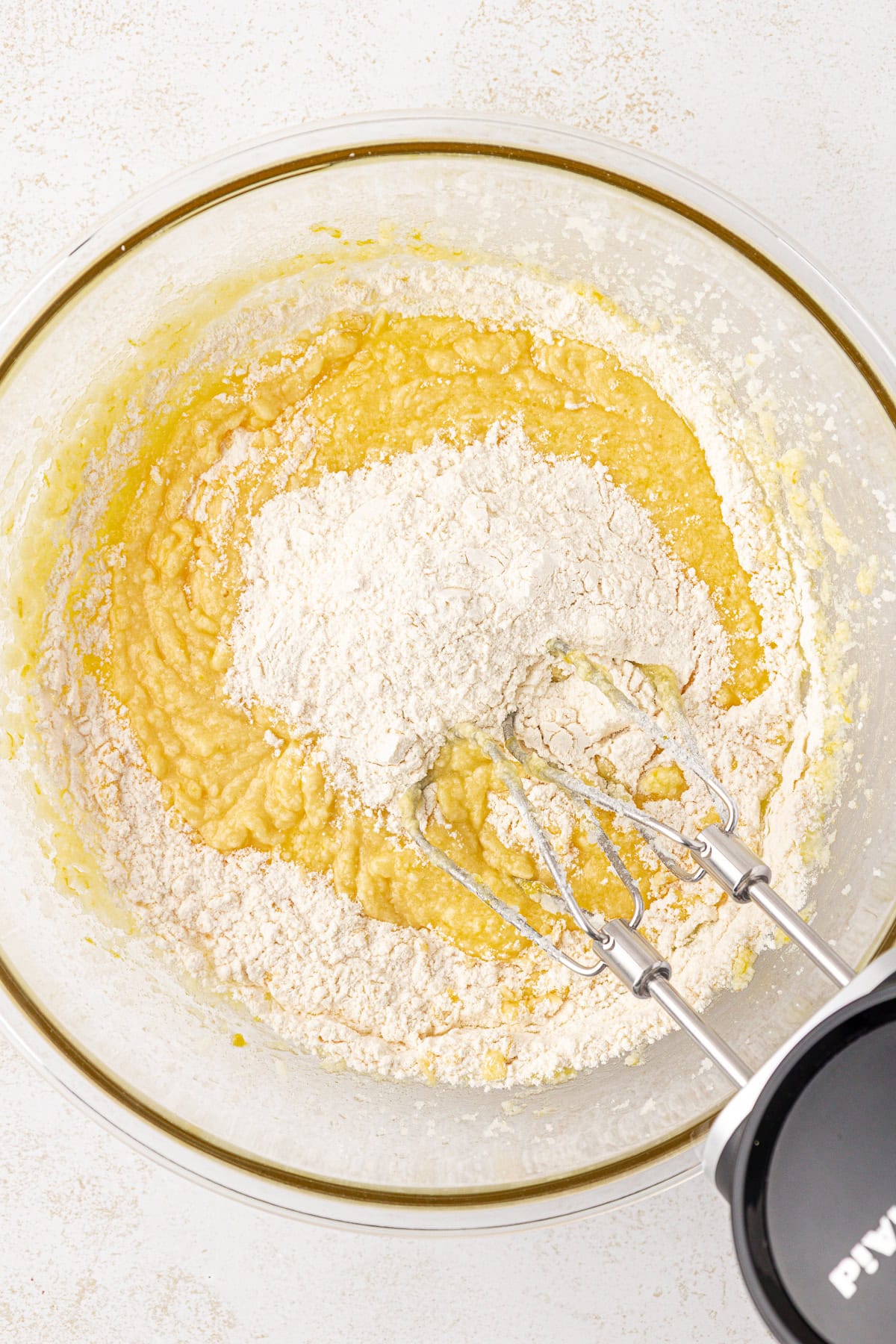 A close-up of a mixing bowl containing cake batter with a pile of flour on top, and an electric hand mixer with beaters partially submerged in the mixture.