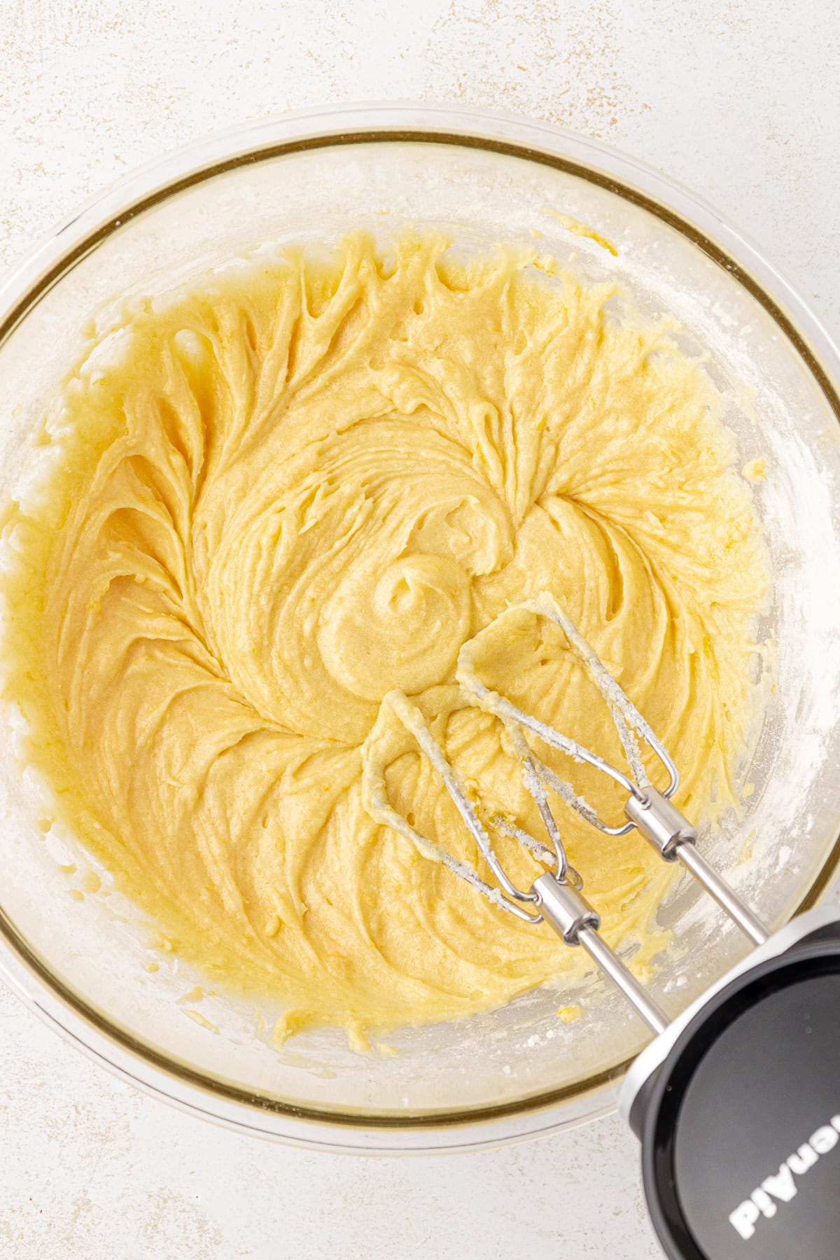 A glass bowl filled with creamy yellow cake batter being mixed with an electric hand mixer; the beaters are resting in the batter.