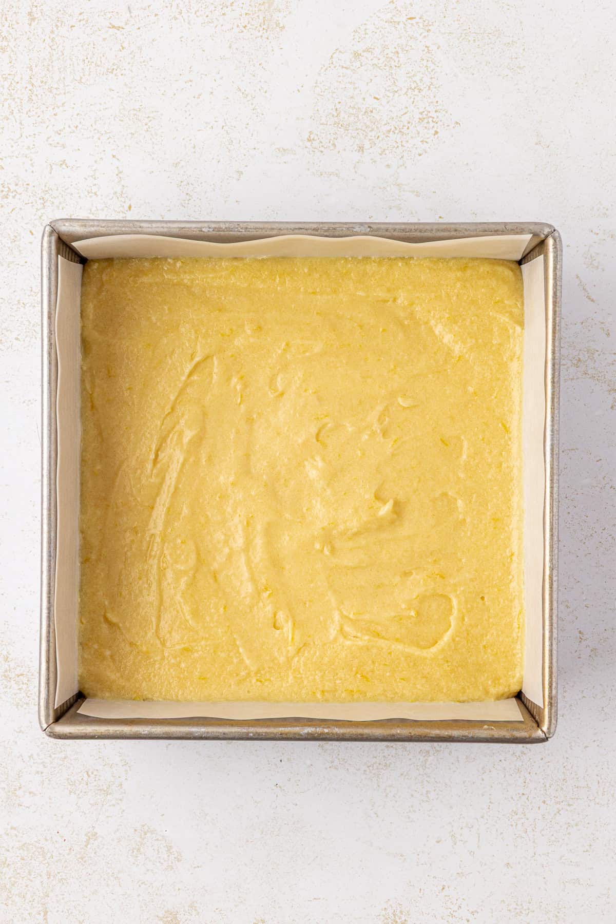 A square baking pan lined with parchment paper is filled with an even layer of smooth, yellow cake batter, ready to be baked.