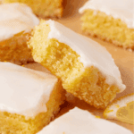 Close-up of lemon brownies with a moist, yellow crumb and a thick layer of white icing on top, arranged on parchment paper with a small lemon wedge visible in the corner.